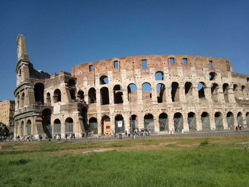 イタリア・ローマの歴史地区にある古代ローマ時代の円形闘技場「コロッセオ」の外観。青空の下、階層状に並ぶ連続したアーチ構造の石造り外壁と、古代の佇まいを残す巨観が捉えられている。