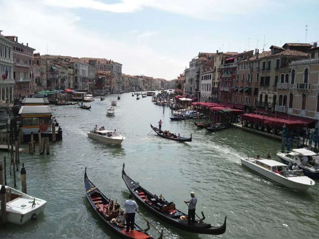 イタリアの世界遺産「ヴェネツィアとその潟」の象徴である大運河(カナル・グランデ)の風景。手前には観光客を乗せたゴンドラが行き交い、水上バスや他の小型ボートも航行している。運河沿いには歴史ある古い建物が立ち並び、活気あるヴェネツィアの日常が捉えられている。