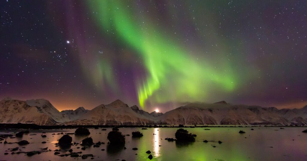 ノルウェー北部の夜空に広がる緑と紫のオーロラ。雪山と水面に映る光